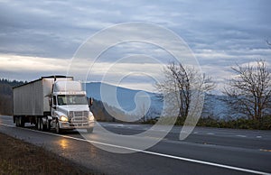White big rig semi truck transporting bulk covered semi trailer driving on the evening wet raining road with turned on headlight