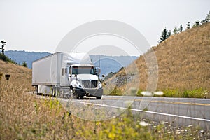 White big rig semi truck transporting goods in dry van semi trailer going on spectacular winding road between the hills with