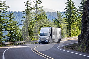 White big rig semi truck transporting goods in refrigerated semi trailer turning on the mountain road with rock wall