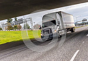 White big rig semi truck transporting industrial cargo on dry van semi trailer running under the bridge across the wide highway