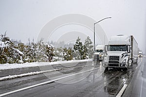 White big rig semi trucks with turned on headlight transporting cargo in semi trailer running on the winter highway road with wet