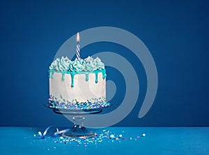 White birthday drip cake with teal ganache and lit candle over dark blue background