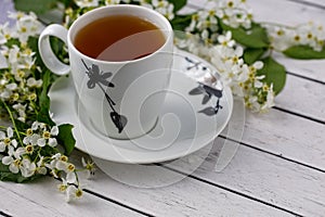White and black cup of black or herbal tea on saucer with white bird cherry tree flowers on white wooden table
