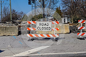 A white, black and orange Road Closed sign at a railroad crossing at an intersection with lush green and bare winter trees