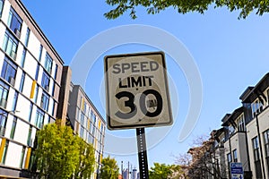 A white and black `Speed Limit 30` sign surrounded by lush green trees, apartment buildings and blue sky in downtown Atlanta