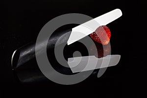 A white blade knife cuts a fresh strawberry and its reflection with black background