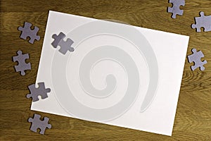 White blank sheet of paper surrounded by puzzle pieces on wooden background. Top view. Copy space for text