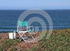 White and blue folding chair with a box of paints is set up by the beach by an artist.