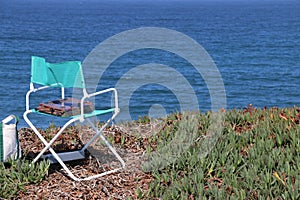 White and blue folding chair with a box of paints is set up by the beach by an artist.