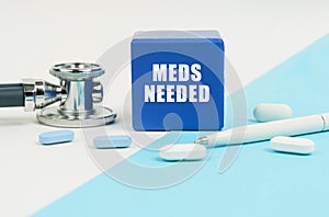 On a white and blue surface are pills, a stethoscope, a pen and a cube with the inscription - Meds Needed