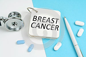 On a white and blue surface are pills, a stethoscope, a pen and a notepad with the inscription - BREAST CANCER