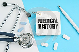 On a white and blue surface are pills, a stethoscope, a pen and a notepad with the inscription - MEDICAL HISTORY