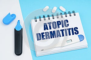 On a white-blue surface are pills, a thermometer, a marker and a notebook with the inscription - ATOPIC DERMATITIS