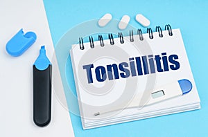On a white-blue surface are pills, a thermometer, a marker and a notebook with the inscription - Tonsillitis