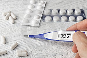 White-blue thermometer with a high temperature of 39.5 degrees Celsius in hand with pills and medical capsules