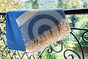 A white and blue Turkish peshtemal / towel on a wrought iron railings with blurry nature in the background.