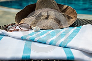 A white and blue Turkish towel, sunglasses and straw hat on rattan lounger with a blue swimming pool as background.