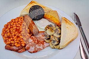 White board with full English breakfast with bacon, fried egg, beans, tomato, roasted sausage, black pudding, scons, hash browns