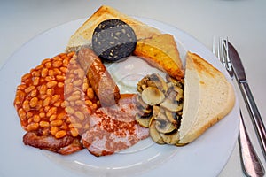 White board with full English breakfast with bacon, fried egg, beans, tomato, roasted sausage, black pudding, scons, hash browns