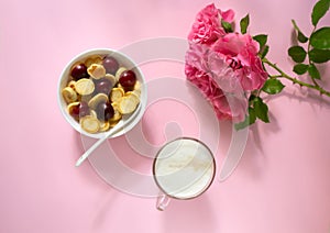 A white bowl of cereal with a tiny pancake with cherries, a cup of cappuccino and flowers on a pink background. Top view.