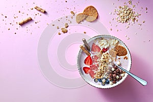 A white bowl of organic yogurt smoothie with strawberries, banana, blueberry, oat flakes and biscuits on pink background