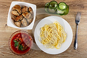 White bowl with pieces fried pork meat, pepper and salt, bowl with lecho, plate with spaghetti, fork on table. Top view