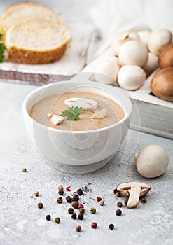 White bowl plate of creamy chestnut champignon mushroom soup on white kitchen background and box of raw mushrooms and fresh bread