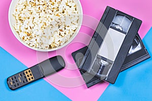 White bowl of popcorn, 2 VHS tapes and old fashioned remote control on a bright pink and blue background.