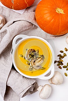 White Bowl of Tasty Pumpkin and Mushrooms Soup with Cream and Pumpkin Seeds on Linen Napkin White Wooden Background Top View