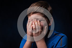 White boy with light brown hair in a blue sports t-shirt covering his face with two hands on  isolated background