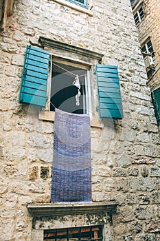 A white bra is drying on a rope on a window with wooden shutters. A stone wall with an open window and a brassiere.