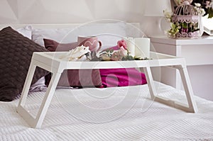 A white breakfast table on a white bed with a cup, flowers and candles on it. Nearby are pink and brown pillows