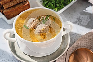 White broth bowl with handles with delicious meatball soup with croutons and herbs on a wooden background, close-up