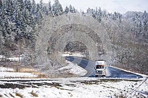 White and brown big rig semi truck transporting cargo in bulk semi trailer running on the winter snow winding road