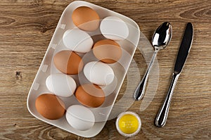 White and brown chicken eggs in plate, spoon, knife, salt shaker on table. Top view