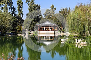 A white and brown Chinese pavilion in the garden with people walking through the pavilion near a deep green lake with lush green t