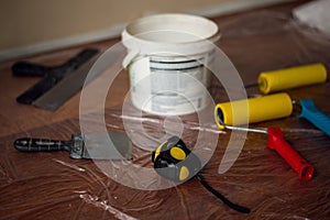 White bucket with paint tape measure construction spatulas and wallpaper rollers on the background of oilcloth on the floor. Room