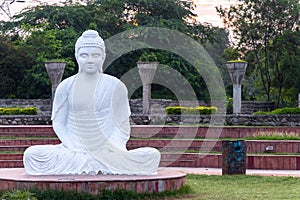 A white Buddha statue in meditation and defocused against  green trees of the park