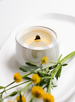 White burning candle with wooden wick and yellow flowers on white tray