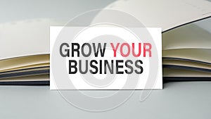 A white business card with grow your business text stands on a desk against the background of an open diary. Unfocused