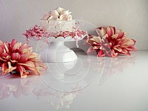 White cake with a frosting flower on top sitting on a white raised cake platter with pink paper shreds decorating it along with