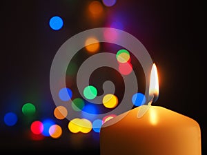A white candle is burning with colorful Christmas lights bokeh in the background