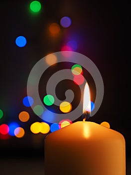 A white candle is burning with colorful Christmas lights bokeh in the background