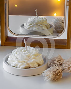 White candle in the shape of a flower on the table near the mirror with reflection and lights in the background. Cozy photo