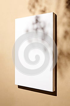 White canvas, blank picture mockup hanging on beige wall with dark shadows of leaves. Poster mockup, empty canvas with shadows of