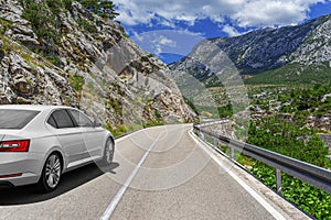 A white car drives along the highway against the backdrop of mountains on a sunny day.