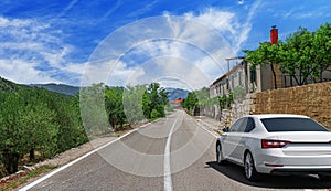 A white car drives along the highway against the backdrop of mountains on a sunny day.