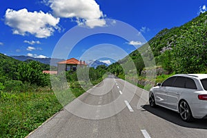 A white car drives along the highway against the backdrop of mountains on a sunny day.