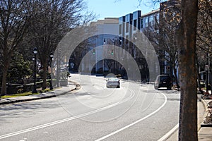 A white car driving along a winding street surrounded by apartment buildings and bare wither trees and parked cars