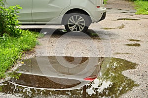 The back of a white passenger car parked at the edge of the road is reflected in a puddle on the sidewalk after rain